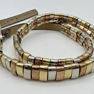 Puede incluir: Un cinturón multicolor con eslabones metálicos rectangulares en tonos dorados, plateados y cobrizos. El cinturón tiene una correa de cuero marrón claro y una hebilla plateada. El cinturón está diseñado para usarse alrededor de la cintura.