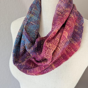 May include: A handwoven infinity scarf in shades of blue, purple, and pink. The scarf is displayed on a white mannequin. The woven fabric has a textured appearance with yellow accents, creating a gradient effect.