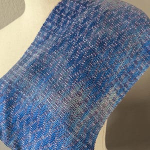 Puede incluir: Bufanda tejida azul y blanca con un patr&oacute;n texturizado. La bufanda est&aacute; drapeada sobre un maniqu&iacute;, mostrando su dise&ntilde;o y variaciones de color. La paleta de colores de la bufanda incluye tonos de azul, blanco y toques de morado.