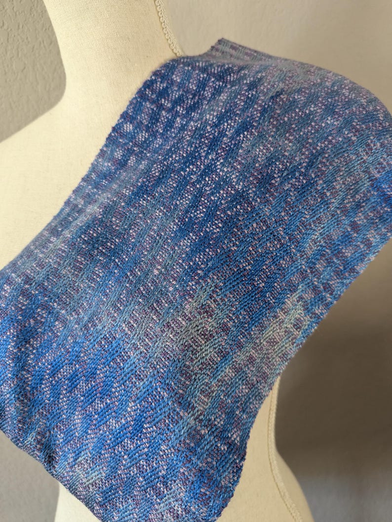 Puede incluir: Bufanda tejida azul y blanca con un patr&oacute;n texturizado. La bufanda est&aacute; drapeada sobre un maniqu&iacute;, mostrando su dise&ntilde;o y variaciones de color. La paleta de colores de la bufanda incluye tonos de azul, blanco y toques de morado.