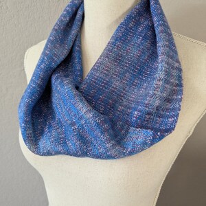 Puede incluir: Un pa&ntilde;uelo infinito azul con una textura tejida, exhibido en un maniqu&iacute;. El pa&ntilde;uelo presenta una mezcla de hilos azules, morados y rosas, creando un patr&oacute;n sutil y texturizado. El accesorio est&aacute; dise&ntilde;ado para usarse alrededor del cuello.