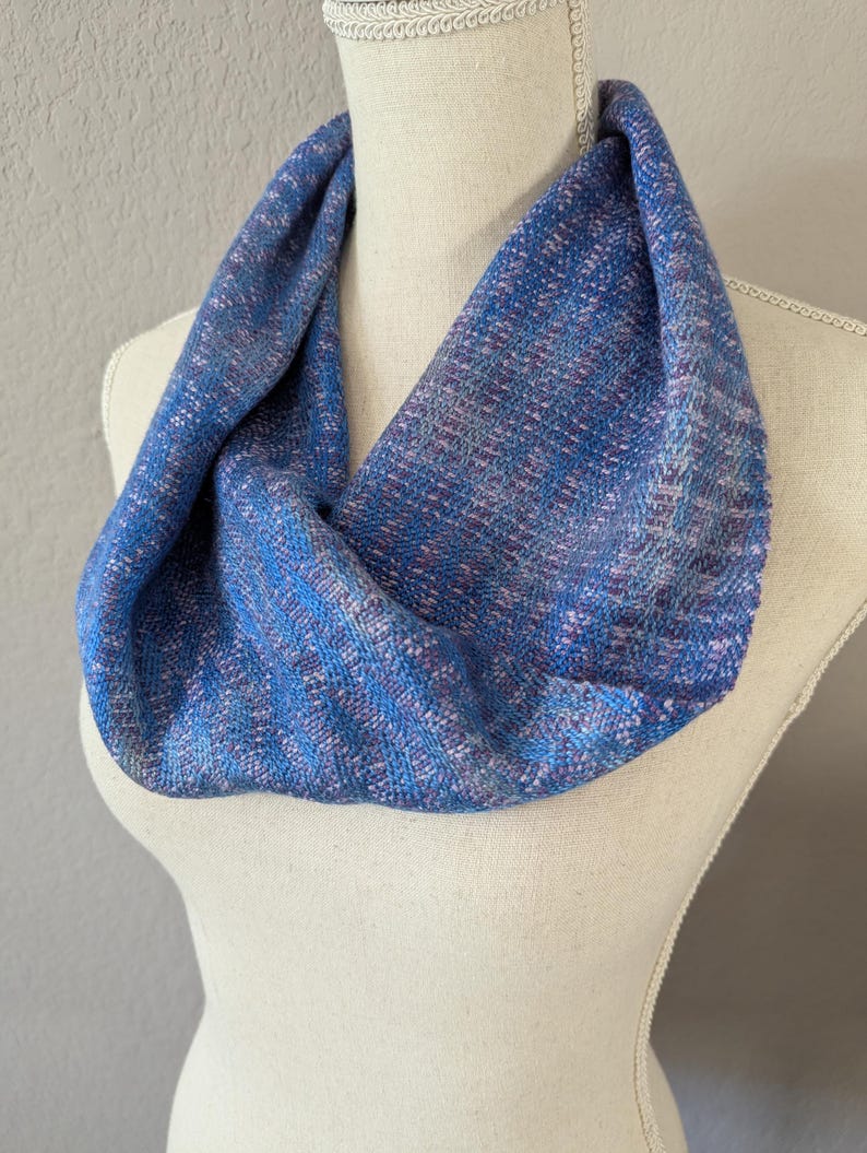 Puede incluir: Un pa&ntilde;uelo infinito azul con una textura tejida, exhibido en un maniqu&iacute;. El pa&ntilde;uelo presenta una mezcla de hilos azules, morados y rosas, creando un patr&oacute;n sutil y texturizado. El accesorio est&aacute; dise&ntilde;ado para usarse alrededor del cuello.