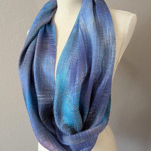 以下が含まれることがあります： 織り目模様の青いインフィニティスカーフ。 スカーフは青、ティール、紫の色合いが特徴です。 スカーフはマネキンに掛けられています。 ファッションアクセサリーです。