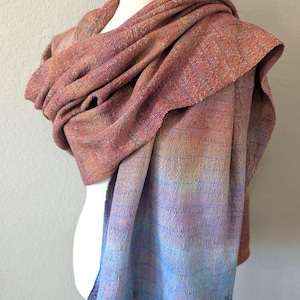 May include: A woven shawl in shades of red, orange, purple, and blue. The shawl is draped over a mannequin, showcasing its gradient colour scheme. The fabric appears soft and textured, with a subtle sheen.