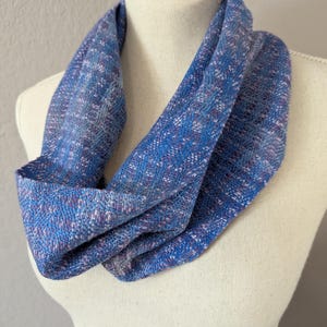 Puede incluir: Bufanda infinita azul con textura tejida. La bufanda presenta una mezcla de hilos azules, morados y blancos, creando un patr&oacute;n sutil. La bufanda est&aacute; drapeada sobre un maniqu&iacute;, mostrando su forma y dise&ntilde;o.