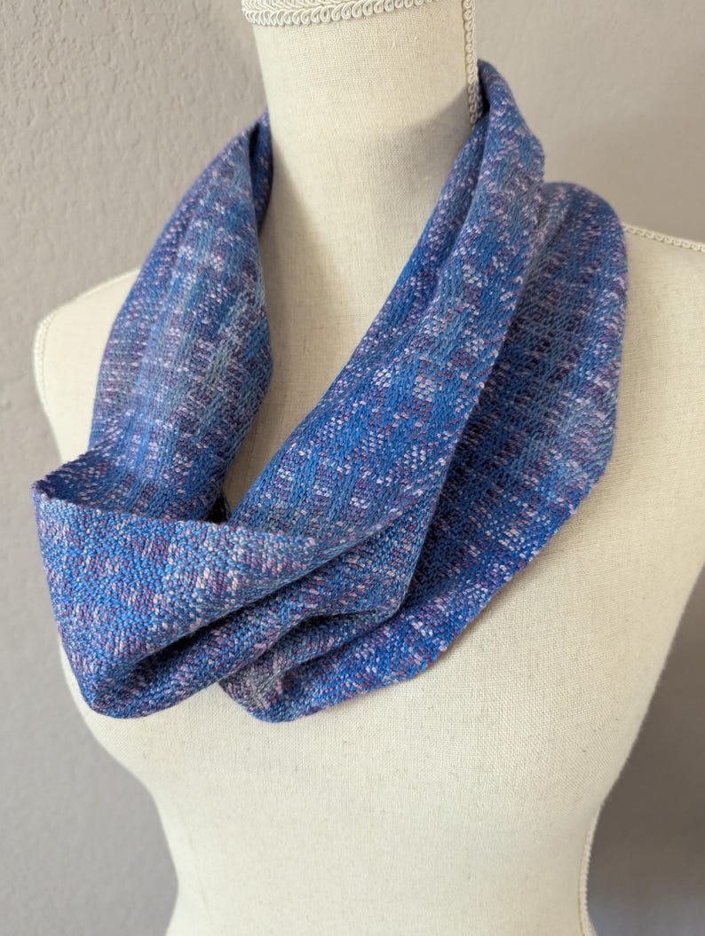 Puede incluir: Bufanda infinita azul con textura tejida. La bufanda presenta una mezcla de hilos azules, morados y blancos, creando un patr&oacute;n sutil. La bufanda est&aacute; drapeada sobre un maniqu&iacute;, mostrando su forma y dise&ntilde;o.