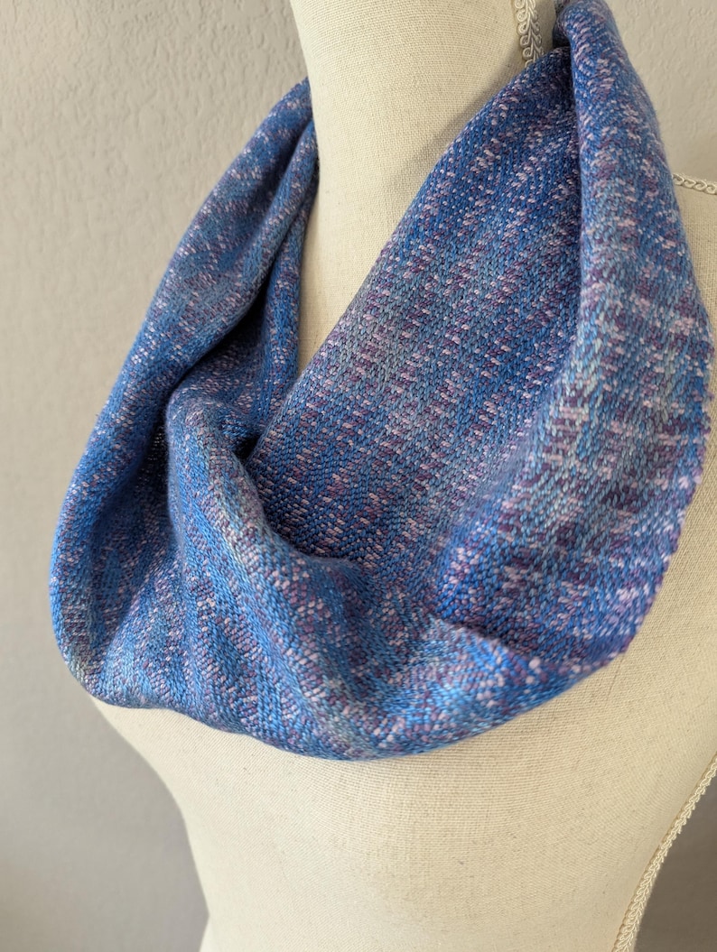 Puede incluir: Bufanda infinita azul y morada sobre un maniqu&iacute;. La bufanda es de tejido con aspecto texturizado. La paleta de colores incluye tonos de azul, morado y blanco. La bufanda es un bucle continuo, dise&ntilde;ada para llevar alrededor del cuello.