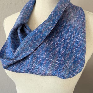 Puede incluir: Un pa&ntilde;uelo infinito azul con una textura tejida, exhibido en un maniqu&iacute;. El pa&ntilde;uelo presenta una mezcla de hilos azules, morados y blancos, creando un patr&oacute;n sutil. La tela est&aacute; drapeada para mostrar su forma y textura.