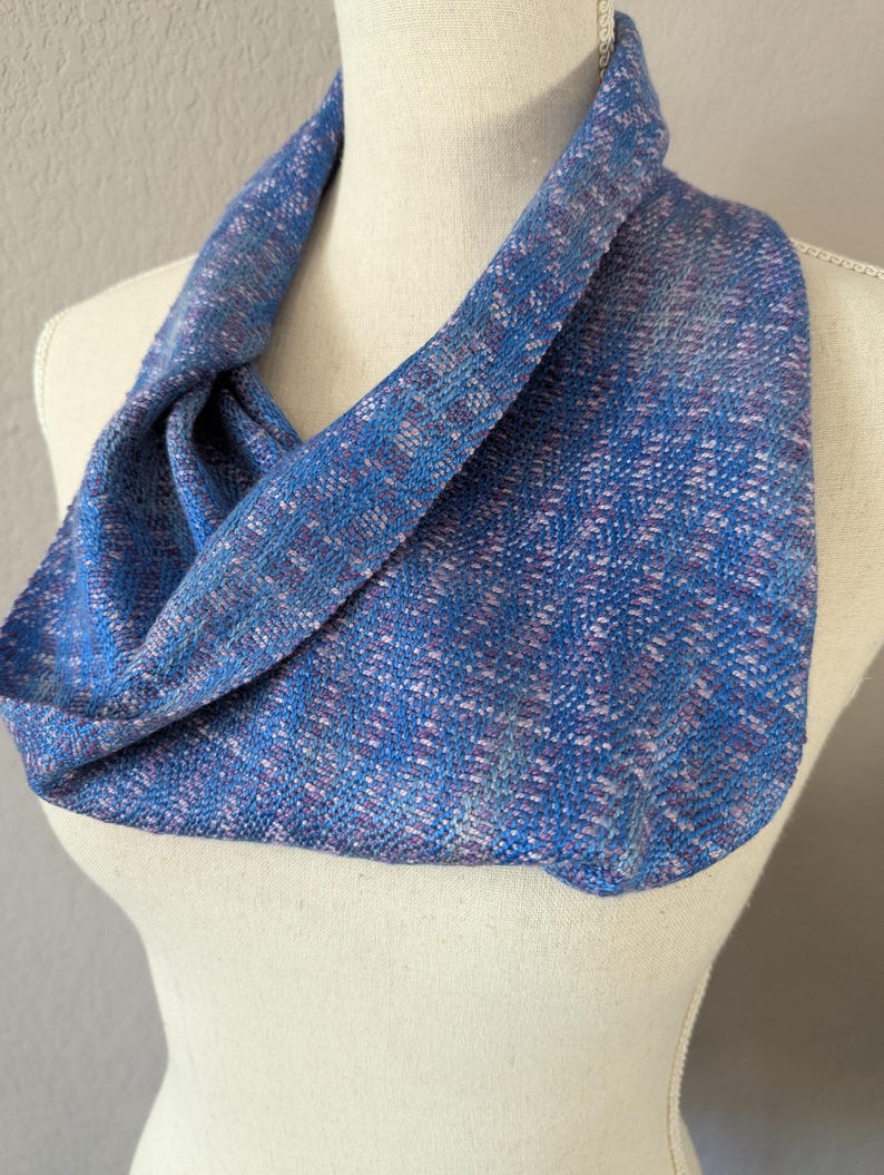 Puede incluir: Un pa&ntilde;uelo infinito azul con una textura tejida, exhibido en un maniqu&iacute;. El pa&ntilde;uelo presenta una mezcla de hilos azules, morados y blancos, creando un patr&oacute;n sutil. La tela est&aacute; drapeada para mostrar su forma y textura.