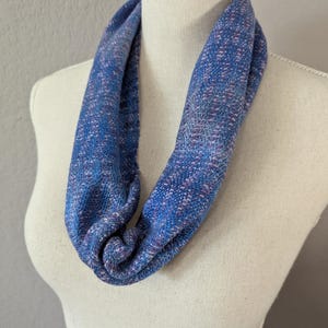 Puede incluir: Bufanda infinita azul con textura tejida, exhibida en un maniqu&iacute;. La bufanda presenta una mezcla de hilos azules y morados, creando un patr&oacute;n sutil. La tela parece suave y c&oacute;moda, adecuada para climas m&aacute;s frescos.