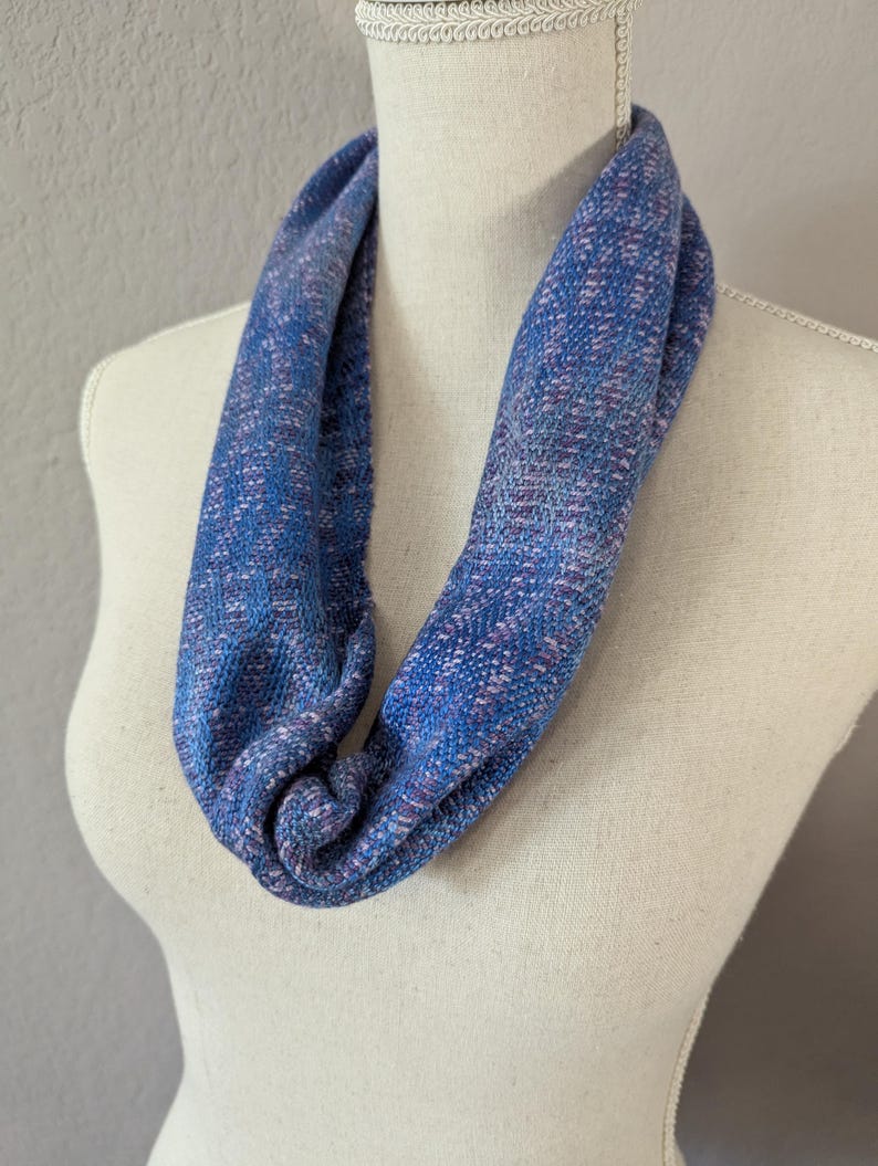 Puede incluir: Bufanda infinita azul con textura tejida, exhibida en un maniqu&iacute;. La bufanda presenta una mezcla de hilos azules y morados, creando un patr&oacute;n sutil. La tela parece suave y c&oacute;moda, adecuada para climas m&aacute;s frescos.
