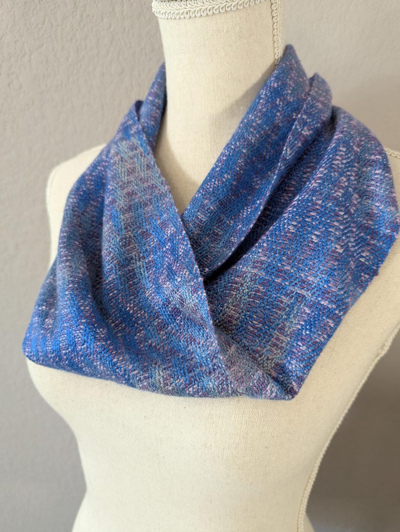 Puede incluir: Un pa&ntilde;uelo infinito azul con una textura tejida, exhibido en un maniqu&iacute;. El pa&ntilde;uelo presenta tonos de azul, morado y blanco, creando un patr&oacute;n texturizado. Este accesorio es una adici&oacute;n elegante a cualquier guardarropa.