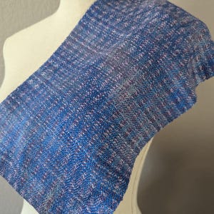 Puede incluir: Una bufanda tejida azul y morada drapeada sobre un maniqu&iacute;. La bufanda tiene un aspecto texturizado con una mezcla de hilos azules, morados y blancos. La tela est&aacute; doblada, mostrando el patr&oacute;n y la textura del material.