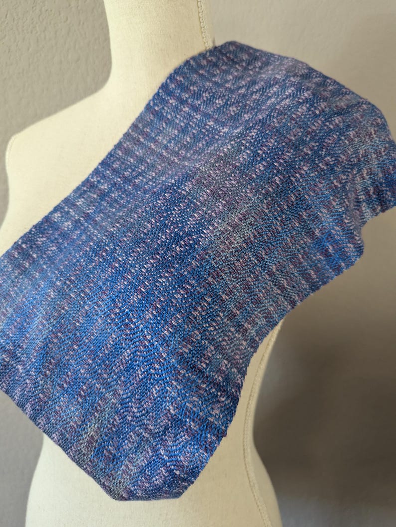 Puede incluir: Una bufanda tejida azul y morada drapeada sobre un maniqu&iacute;. La bufanda tiene un aspecto texturizado con una mezcla de hilos azules, morados y blancos. La tela est&aacute; doblada, mostrando el patr&oacute;n y la textura del material.