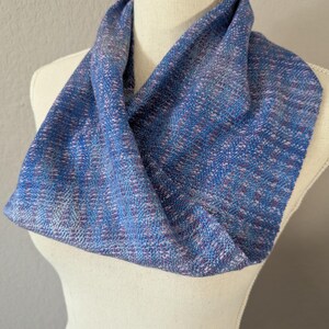 Puede incluir: Bufanda infinita azul y morada exhibida en un maniqu&iacute;. La bufanda tiene una textura tejida con hilos azules, morados y blancos. La tela est&aacute; drapeada en forma circular, mostrando las variaciones de color y la textura. Accesorio de moda.