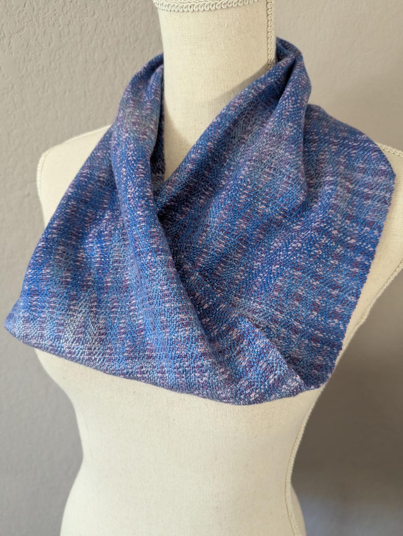 Puede incluir: Bufanda infinita azul y morada exhibida en un maniqu&iacute;. La bufanda tiene una textura tejida con hilos azules, morados y blancos. La tela est&aacute; drapeada en forma circular, mostrando las variaciones de color y la textura. Accesorio de moda.