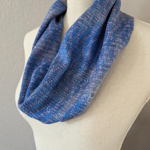 Puede incluir: Bufanda infinita azul con textura tejida, exhibida en un maniqu&iacute;. La bufanda presenta tonos de azul, con toques de morado y blanco, creando un patr&oacute;n texturizado. La tela parece suave y ligera, adecuada para varias estaciones.