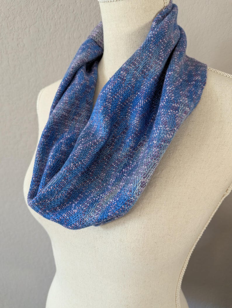 Puede incluir: Bufanda infinita azul con textura tejida, exhibida en un maniqu&iacute;. La bufanda presenta tonos de azul, con toques de morado y blanco, creando un patr&oacute;n texturizado. La tela parece suave y ligera, adecuada para varias estaciones.