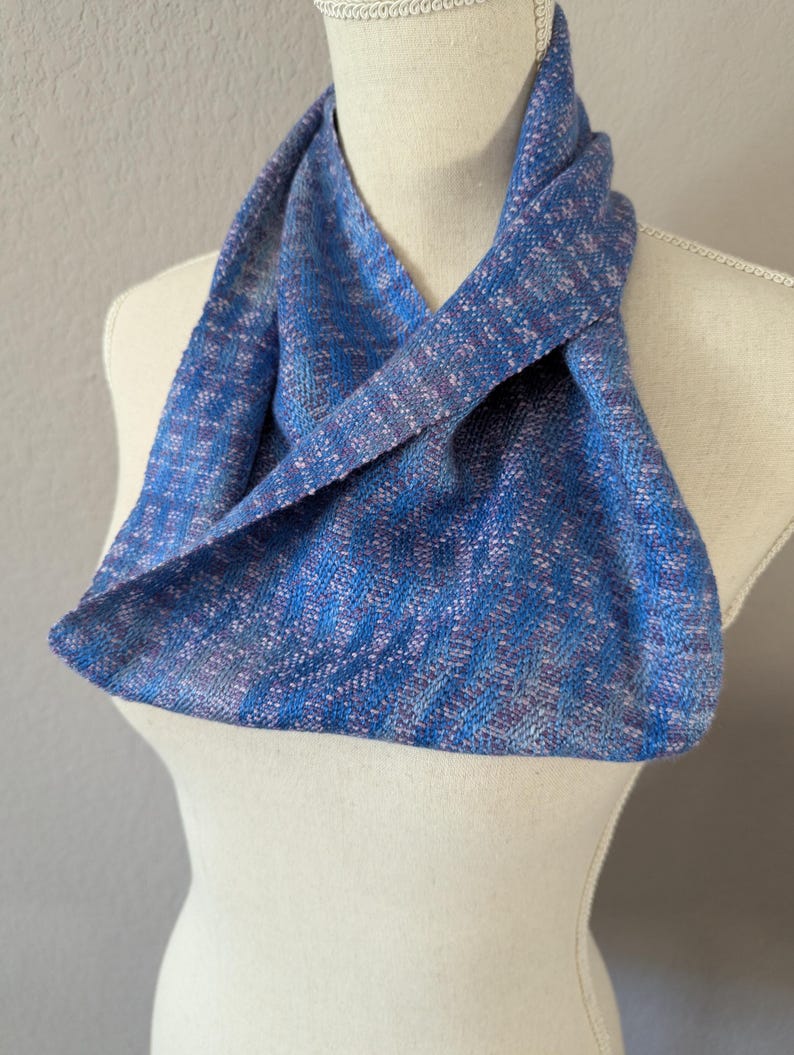 Puede incluir: Bufanda infinita azul con un patr&oacute;n tejido. La bufanda se muestra en un maniqu&iacute; y presenta tonos de azul, morado y blanco. La tela parece suave y ligera, adecuada para climas m&aacute;s frescos. Un accesorio vers&aacute;til.