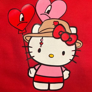 May include: A red hoodie featuring a white Hello Kitty illustration wearing a brown hat with a red bow. Hello Kitty is holding two red and pink heart-shaped balloons with sad faces.