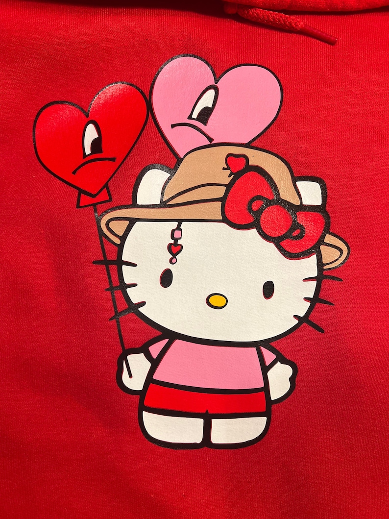 May include: A red hoodie featuring a white Hello Kitty illustration wearing a brown hat with a red bow. Hello Kitty is holding two red and pink heart-shaped balloons with sad faces.