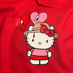 May include: Red hoodie featuring a white Hello Kitty character wearing a brown hat with a red bow. The character is holding two red and pink heart-shaped balloons with sad faces.