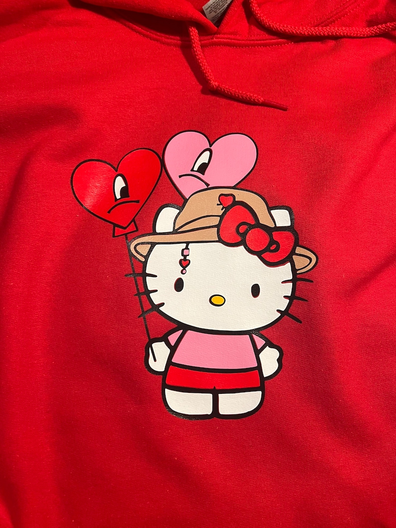 May include: Red hoodie featuring a white Hello Kitty character wearing a brown hat with a red bow. The character is holding two red and pink heart-shaped balloons with sad faces.
