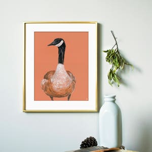 May include: A framed illustration of a brown and white goose with a black head and neck, set against a peach background. The frame is gold and the illustration is hanging on a white wall.