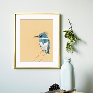 May include: A framed print of a blue and white bird with a long beak, perched on a branch. The print is on a light beige background and is framed in gold.