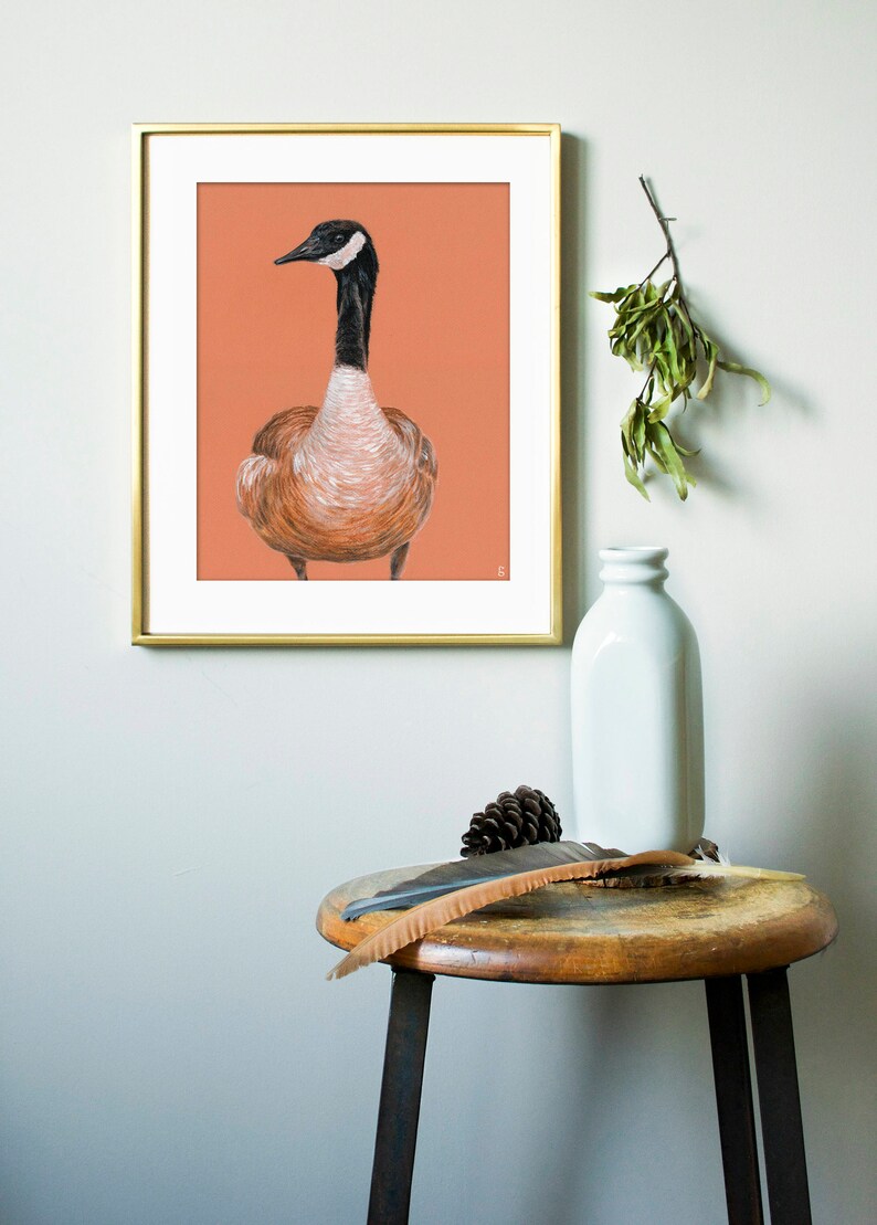 May include: A framed illustration of a brown and white goose with a black head and neck, set against a peach background. The frame is gold and the illustration is hanging on a white wall.