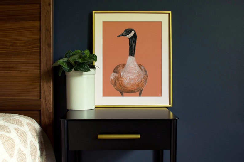 May include: A framed print of a goose with a black head and neck, a white collar, and brown feathers. The print is on a peach background and is framed in gold.