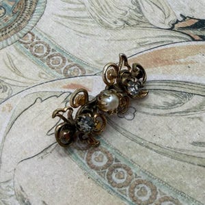 May include: A vintage-style hair clip with an ornate design featuring small clear rhinestones and a central pearl. The clip is gold-toned and rests on a decorative print with a woman in a flowing gown, in shades of cream, teal, and brown.