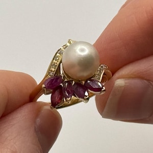 May include: A gold ring featuring a large, round, white pearl at the center, surrounded by smaller, marquise-cut, red gemstones. The ring's band is adorned with small, clear gemstones.