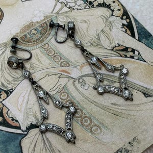 Peut inclure: Boucles d'oreilles à clip argentées antiques avec strass transparents. Les boucles d'oreilles ont un design géométrique avec une forme de diamant centrale et des éléments pendants. Les boucles d'oreilles sont présentées sur une estampe d'art vintage.