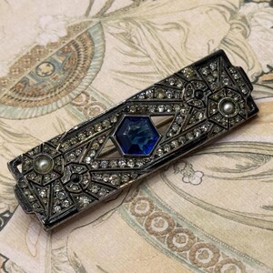 May include: A rectangular vintage brooch with a silver-tone metal frame. It features a central blue gemstone and is encrusted with clear rhinestones. Small pearl accents enhance the antique look. The brooch is set against a patterned background, typical of the era.