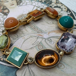 May include: A gold-toned bracelet featuring a variety of colorful gemstones. The bracelet includes orange, green, teal, and purple stones in various shapes, including round, oval, and square. The bracelet is a vintage piece.
