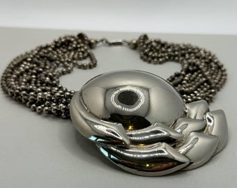 May include: A silver-toned statement necklace featuring multiple strands of small, round beads. The necklace has a large, polished, oval-shaped centerpiece with a floral design on the bottom. The necklace is designed to be worn around the neck.