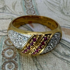 May include: A gold band ring featuring a central diagonal row of small red rubies flanked by two sections of pavé-set clear white diamonds. The ring has a polished gold finish and a wide, tapered design that wraps around the finger.