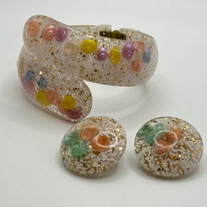 May include: A set of vintage clear plastic jewelry with a gold glitter finish. The bracelet is a wide, curved band with a clasp. The earrings are round and have a similar design to the bracelet. The jewelry is decorated with colorful, small, plastic flowers.