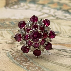 May include: A silver cluster ring with a floral design. The ring is set with numerous round, deep red rubies and small diamonds. The gemstones are arranged in a flower-like pattern, creating a striking visual effect.