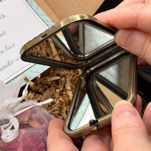May include: An open, antique-style, brass-toned compact mirror with two mirrored surfaces. The compact is held open, revealing the reflective surfaces. The compact is part of a gift set that includes a decorative box and rose and lavender sachets.