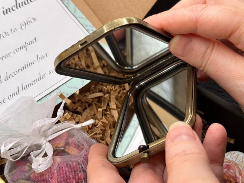 May include: An open, antique-style, brass-toned compact mirror with two mirrored surfaces. The compact is held open, revealing the reflective surfaces. The compact is part of a gift set that includes a decorative box and rose and lavender sachets.