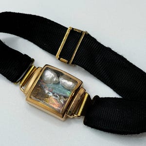 Peut inclure: Un bracelet en tissu noir avec une pièce maîtresse rectangulaire de couleur or. La pièce maîtresse présente une fenêtre rectangulaire transparente avec deux perles blanches et un matériau irisé. Le bracelet a un fermoir doré.