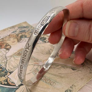 Peut inclure: Un bracelet jonc de couleur argentée polie présentant une série de motifs géométriques gravés et répétitifs sur sa surface extérieure. L'anneau métallique possède une finition lisse et réfléchissante ainsi qu'un profil arrondi.