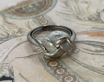 May include: A silver-toned ring with a textured knot design. The ring features a double band and a central knot element, creating a unique and elegant look. The ring is likely made of metal and is suitable for everyday wear or special occasions.