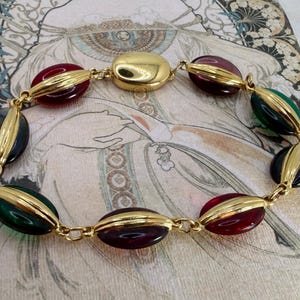 Puede incluir: Un brazalete dorado con cuentas ovaladas en tonos rojo, verde y azul oscuro. Las cuentas est&aacute;n unidas por metal dorado y tienen un acento dorado. El brazalete tiene una est&eacute;tica vintage.