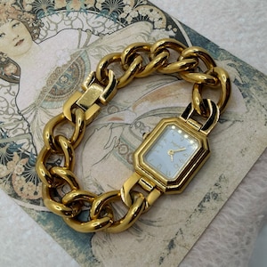 May include: A gold watch with a light blue face and a chunky chain-link bracelet. The watch has a rectangular face with gold hands and hour markers. The bracelet is made of large, rounded links. The watch is displayed on vintage paper.