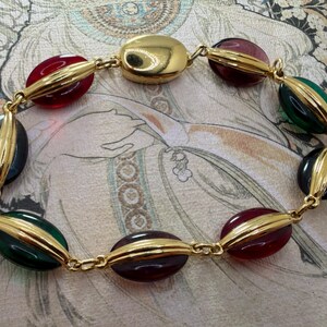 Puede incluir: Un collar dorado con cuentas ovaladas en rojo, verde, negro y morado. Las cuentas est&aacute;n unidas por eslabones dorados. El collar tiene un estilo vintage.