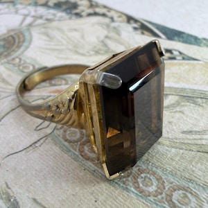 May include: A gold-toned ring featuring a large, rectangular, faceted dark brown gemstone set in a metal frame. The band of the ring displays an embossed floral pattern along the sides, and the stone is secured by four small metal prongs at the corners.
