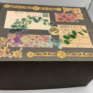 May include: A dark brown box decorated with vintage-style postcards, floral patterns, and gold embellishments. The box features botanical illustrations, including eucalyptus leaves and floral designs, with the word "Post Card" visible.